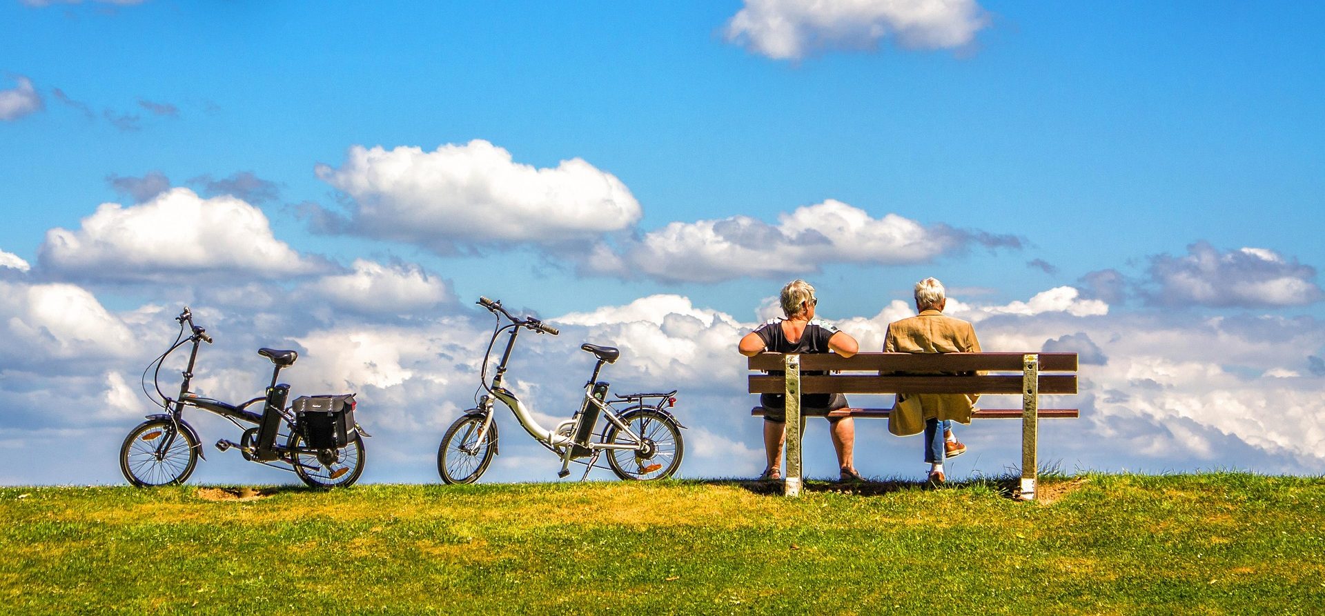 luxury travel specialist Two people sit on a bench with bicycles nearby, under a blue sky with fluffy clouds.