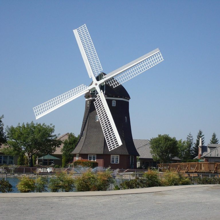 Eco-Friendly Vacations A large windmill beside a calm body of water and trees under a clear blue sky.