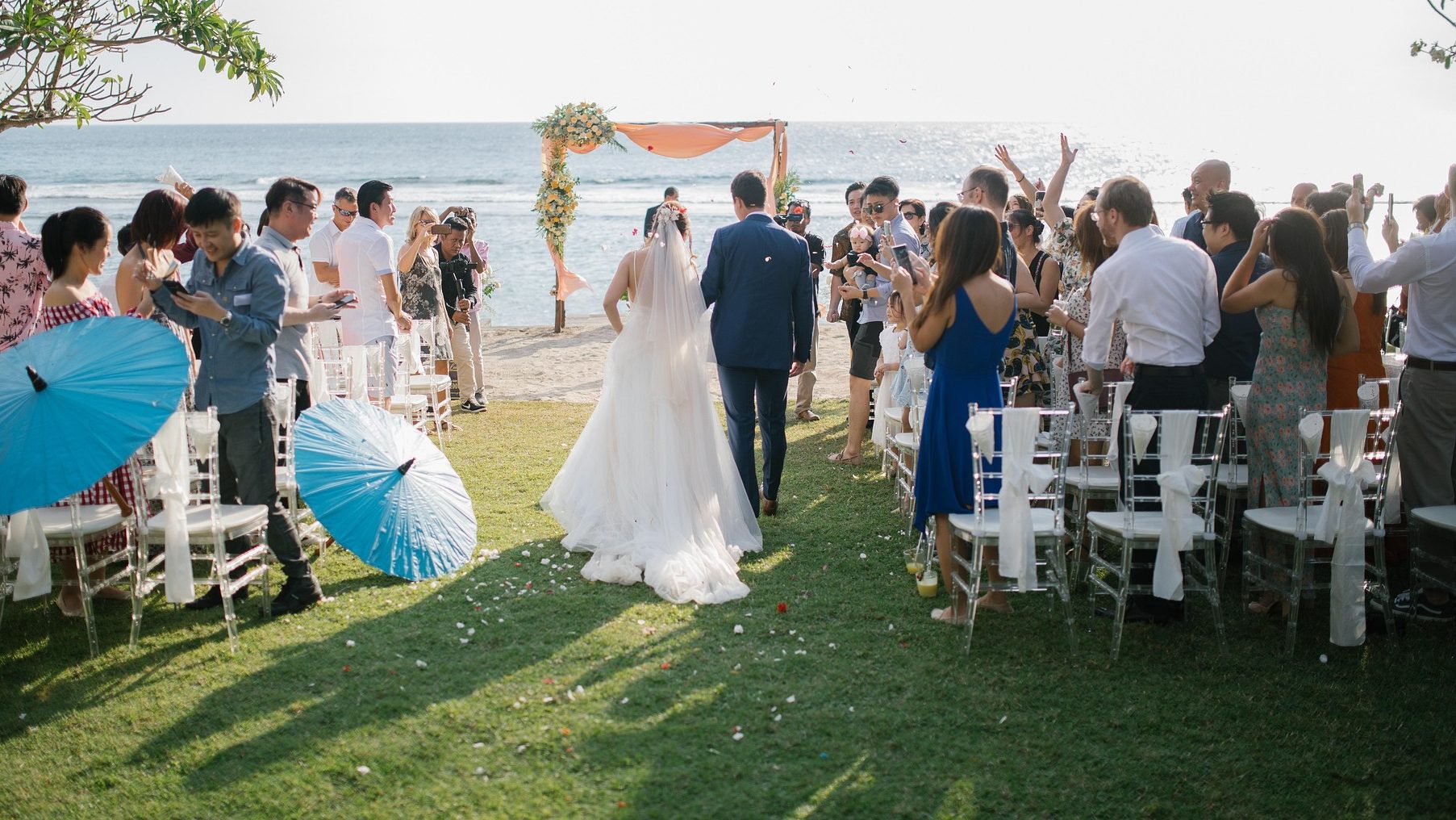 Tailored Travel Experiences Bride and groom walk towards the beach while guests celebrate with umbrellas.