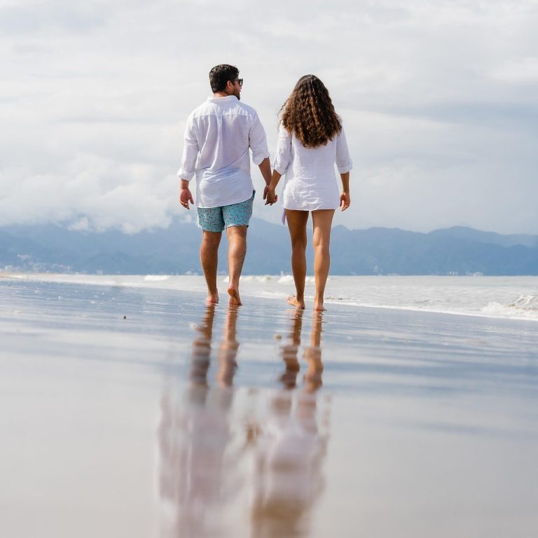 Romantic Vacation Couple walking hand-in-hand along a beach with gentle waves and cloudy sky.